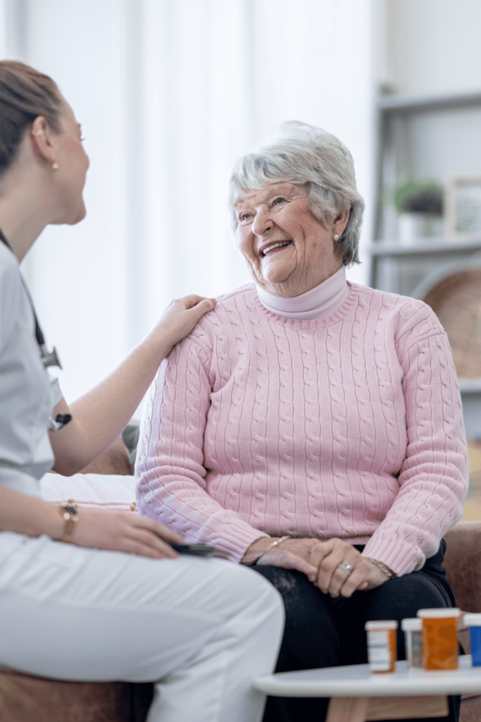 An elderly woman in a pink sweater smiles warmly while sitting and talking with a caregiver in white scrubs who is gently touching her shoulder, creating a caring and supportive atmosphere.
